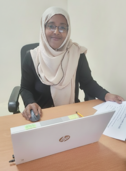 An EYLE student wearing glasses and a white head covering is smiling at the camera as she sits at a laptop.  